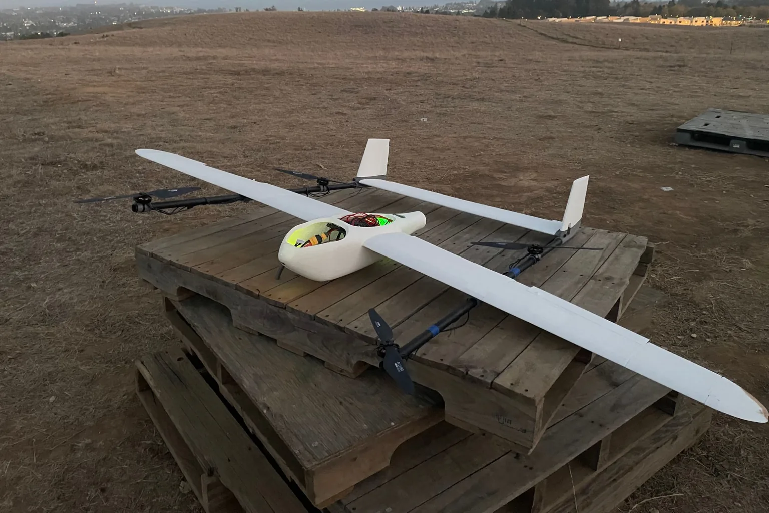 A white fixed-wing VTOL aircraft with four lift rotors on wing booms, resting on a stack of pallets in a dry field at dusk.