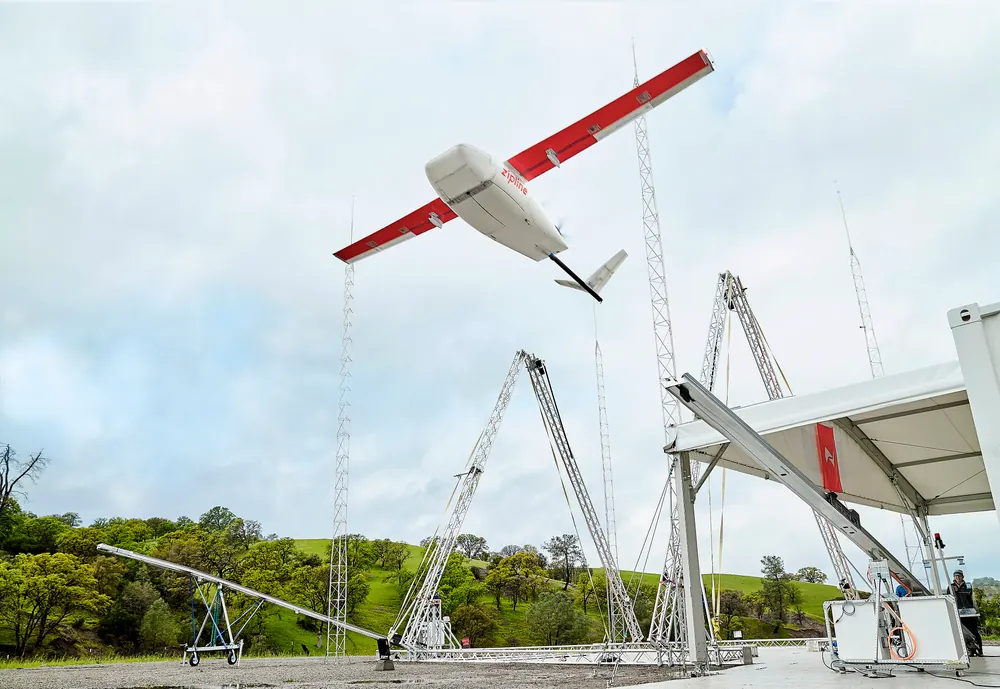 A red and white fixed-wing delivery aircraft leaving a catapult launcher at a rural distribution site.